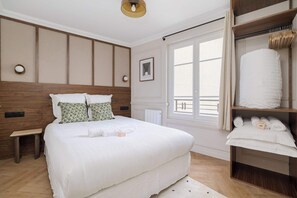 Une chambre bien rangée, avec un grand lit, une table de chevet, un banc, une armoire contenant des serviettes et une fenêtre avec des rideaux.