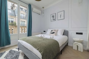 Ein Schlafzimmer mit Bett, einem Fenster mit Vorhängen, einem Heizkörper, einer Wandleuchte und einem kleinen weißen Hocker.