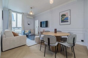Ein modernes Wohnzimmer mit einem runden Holzesstisch, grau gepolsterten Stühlen, einem weißen Sofa, einem Flachbildfernseher und einem gerahmten Bild an der Wand.