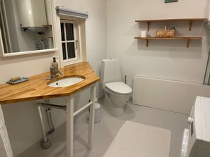 Kylpyhuoneessa on puinen allaskaappi, valkoinen allas, wc ja pieni hylly, jossa on hygieniatuotteita.