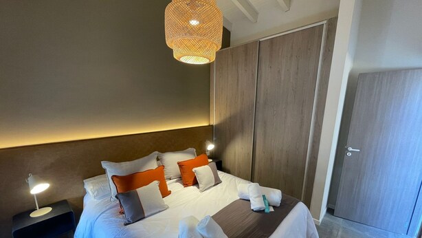 A modern hotel room with a large bed, a bedside table with a lamp, a wardrobe, and a door.