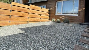 A modern two-story house with a wooden deck, a gravel driveway, and a stone wall.