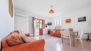 Een ruime woonkamer met een bank, eettafel en stoelen, een open deur naar buiten, en een aan de muur bevestigde televisie.
