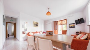 Een eethoek met een houten tafel en witte stoelen, een bank met oranje kussens, en een aan de muur bevestigde televisie.