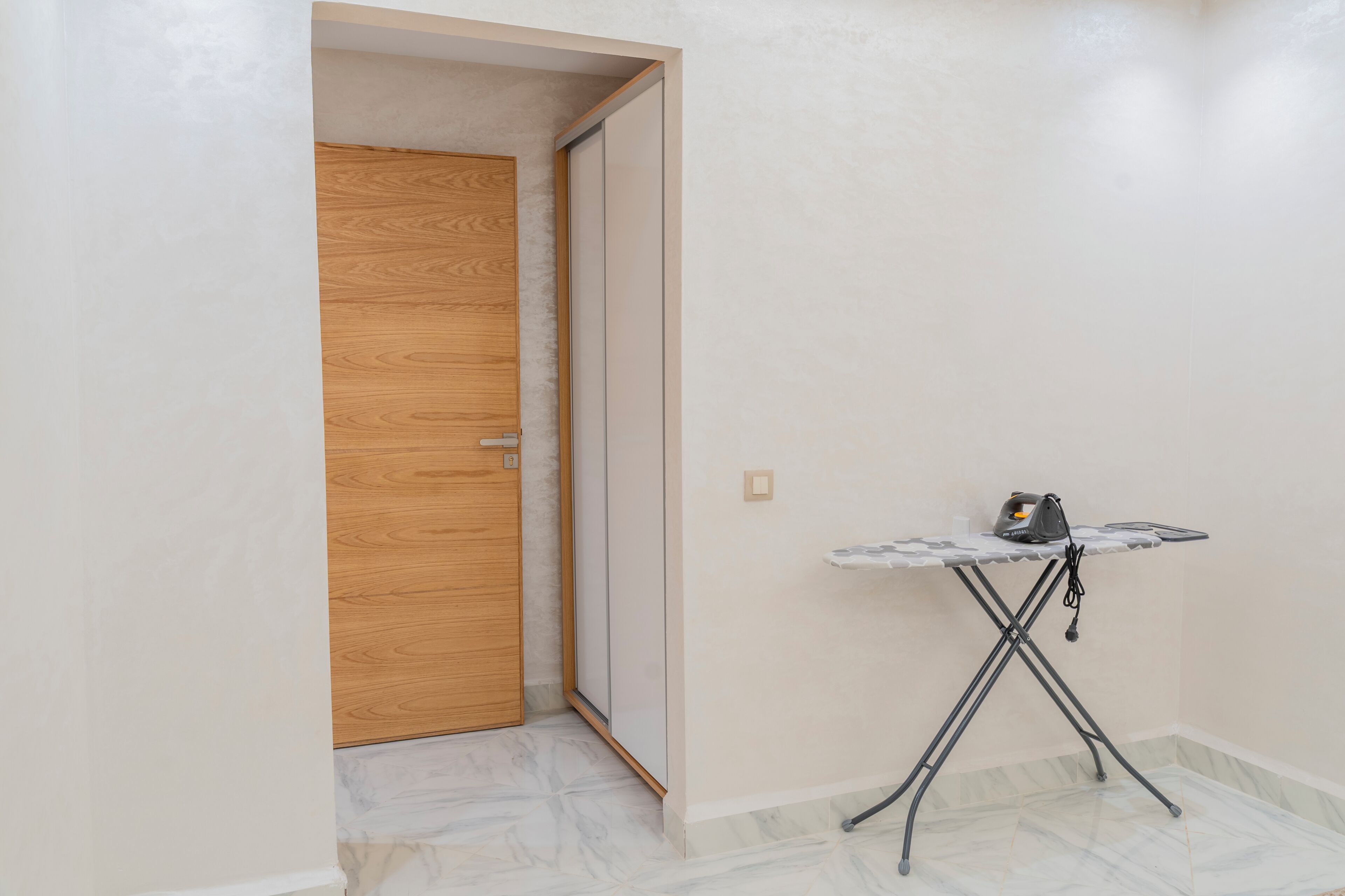 A room with a wooden door, a small ironing station with an iron, and a tiled floor.