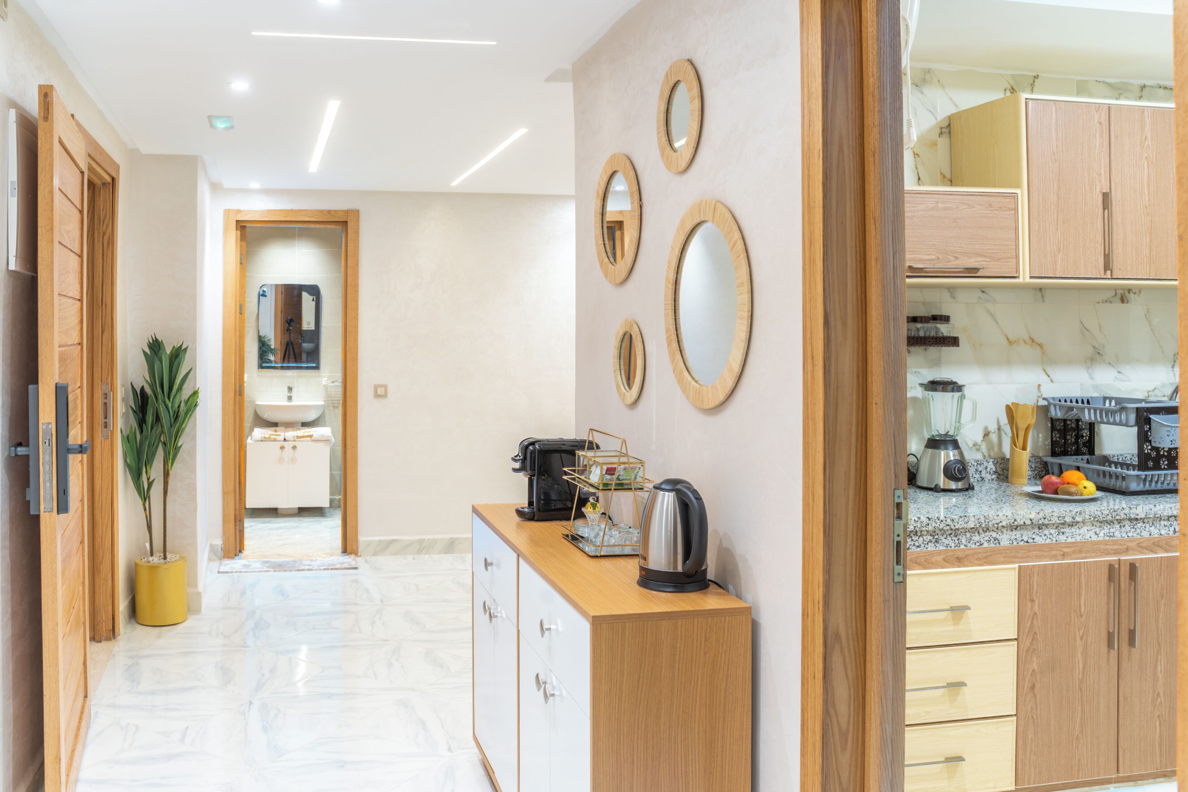 A modern hotel corridor with a wooden reception desk, a potted plant, and a view into a bathroom and kitchen area.