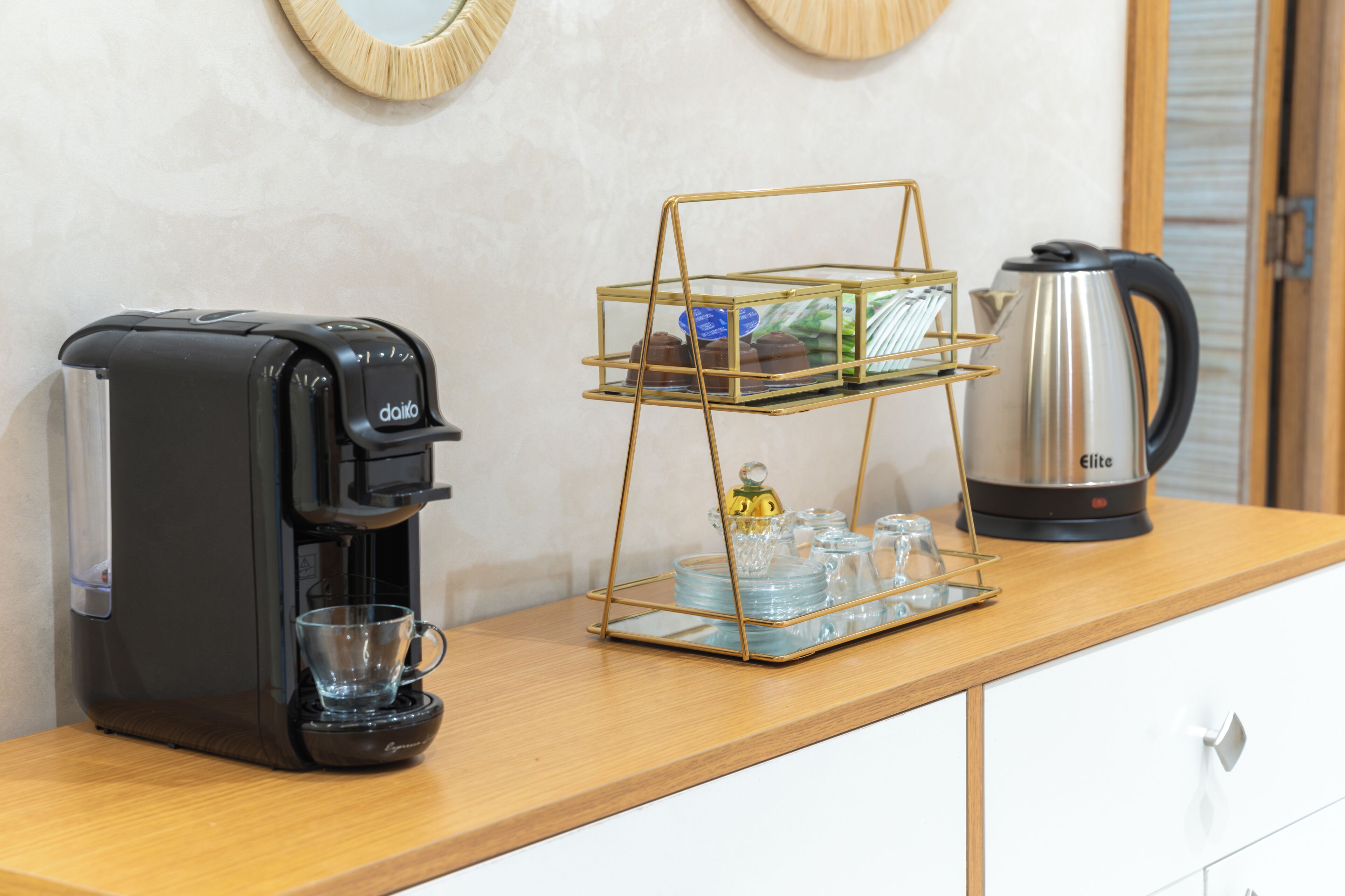 A coffee maker, a glass display stand with items, and a kettle on a wooden counter.