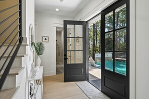 Un interior moderno con una escalera, una puerta de marco negro que da a una zona de piscina y una consola blanca con un jarrón.