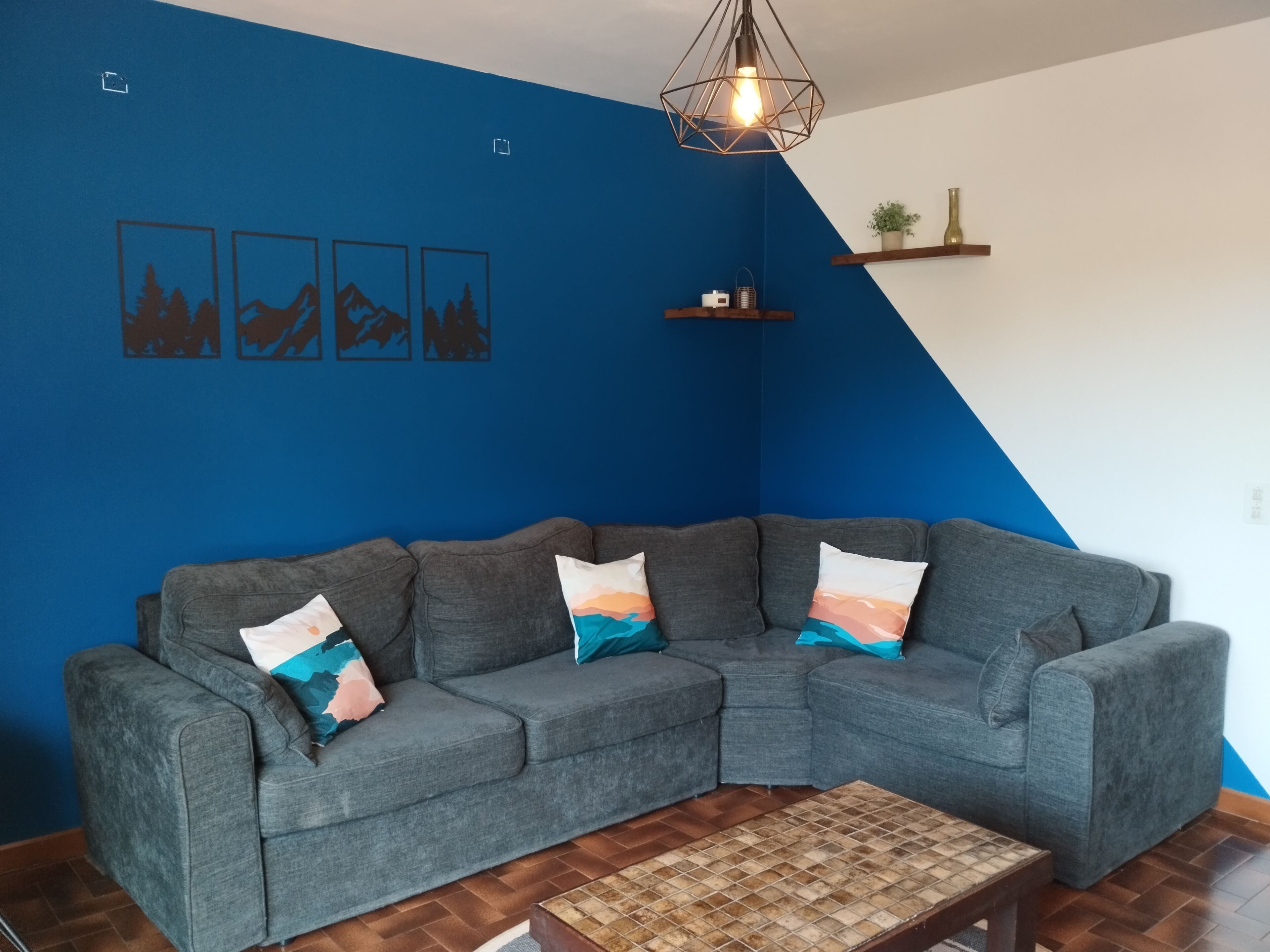 A living room with a blue accent wall, a grey sectional sofa with patterned cushions, a wooden coffee table, and a hanging light fixture.