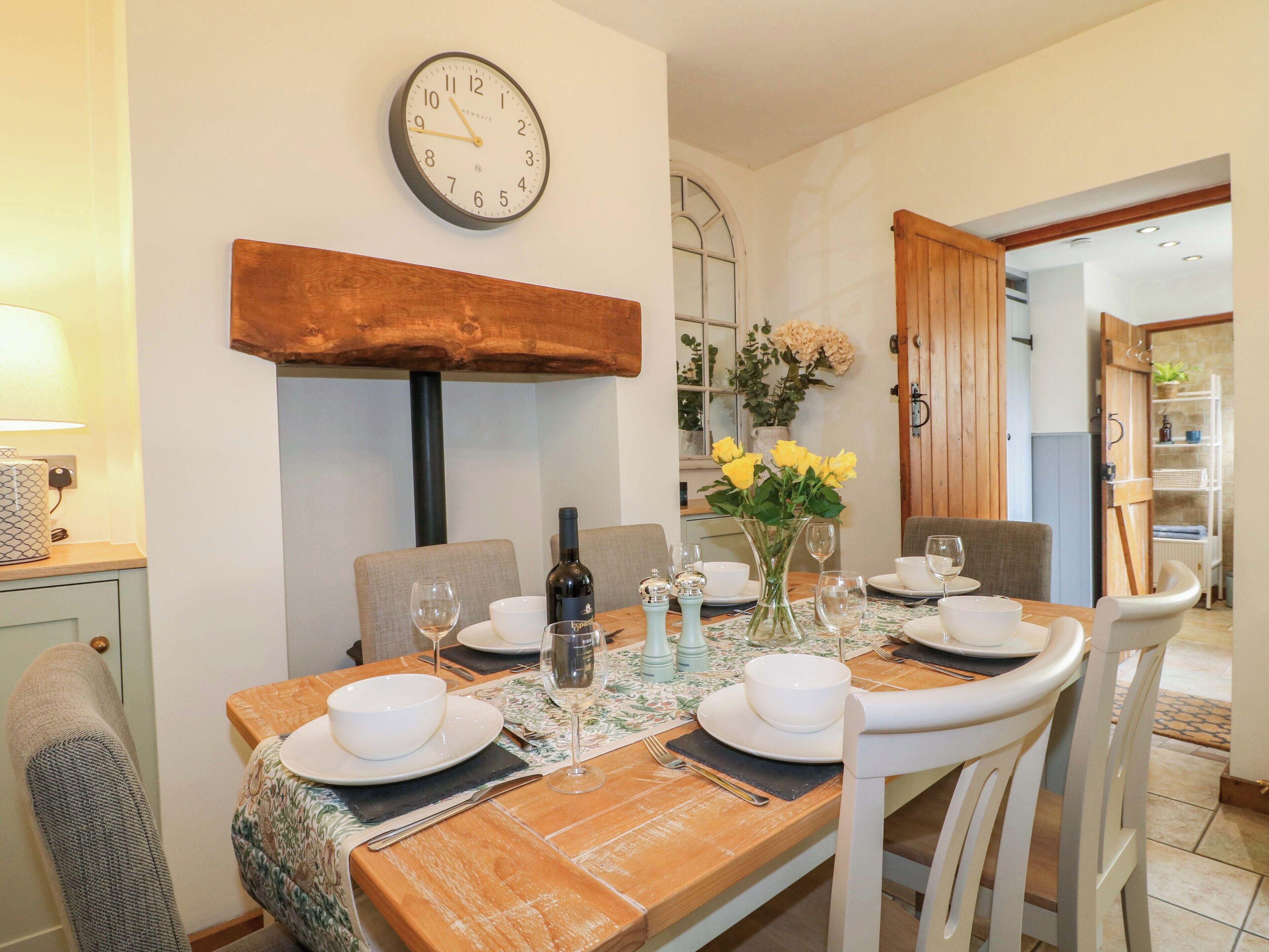 A dining area with a wooden table, white chairs, a clock on the wall, and a vase with yellow flowers.