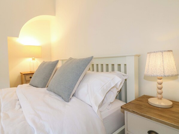 A bed with white bedding, two pillows with a chevron pattern, a wooden nightstand with a lamp, and a white dresser with a mirror. - Lumford Mill Cottage (Bakewell)