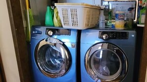 Zona de lavandería con dos lavadoras apiladas, una cesta y productos de limpieza.