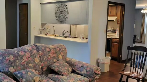 Sala de estar con sofá floral, una silla de madera, una zona de cocina con pileta y muebles bajos, y un adorno de pared.