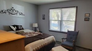 Un dormitorio con cama, una silla, un escritorio, una ventana con persianas y adornos en la pared.