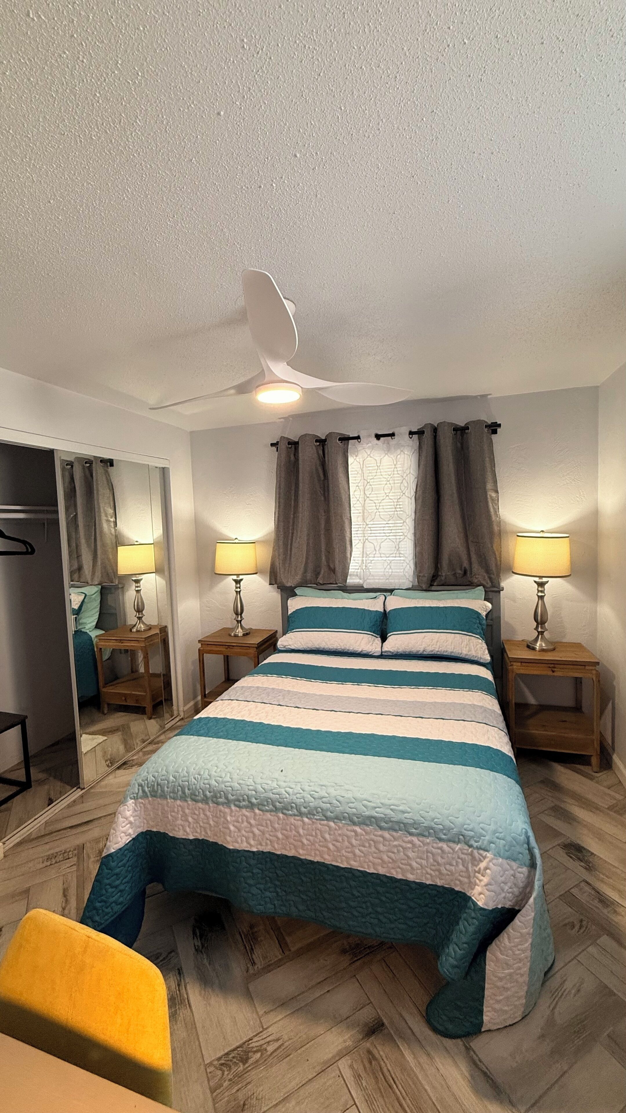 Un dormitorio con una cama, dos mesitas de noche con lámparas, un ventilador de techo, una ventana con cortinas y un espejo.