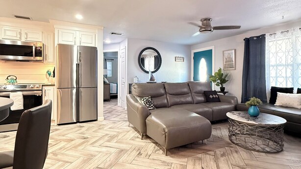 A modern living room with a herringbone floor, a sectional sofa, a coffee table, and a kitchen area with stainless steel appliances.