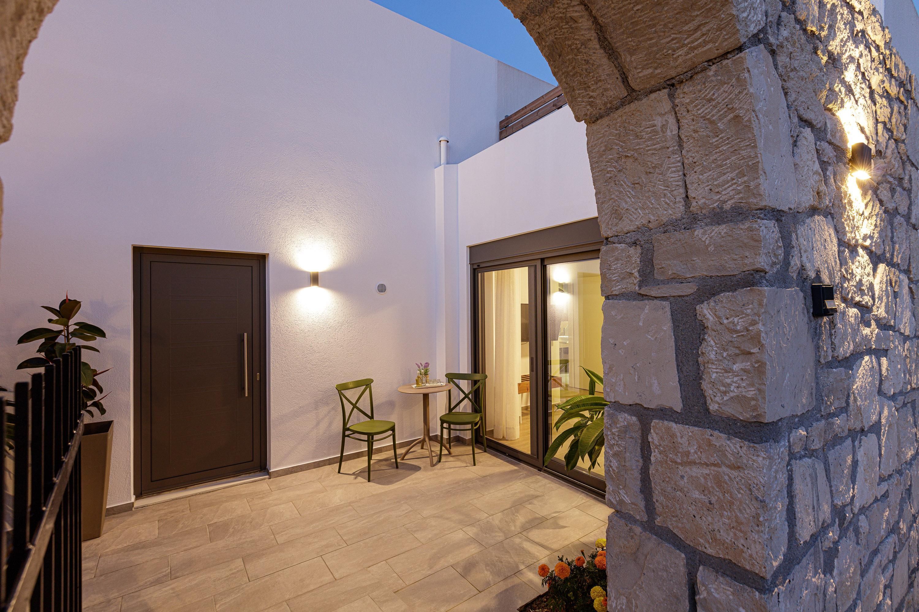Uma área de pátio com porta, duas cadeiras, uma mesa pequena e um muro de pedra com iluminação.