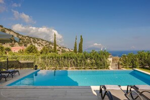 A swimming pool with lounge chairs, a view of the sea, and a hilly landscape. - Petradi Villa Skala II (Kefalonia)