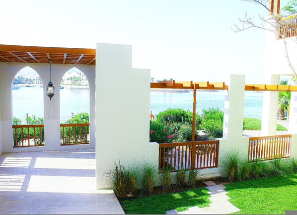 A covered outdoor area with a view of a body of water, a pergola, and a pathway leading to a white building.