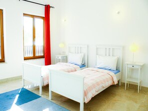 A double room with two beds, a window with a red curtain, bedside lamps, and a blue rug.