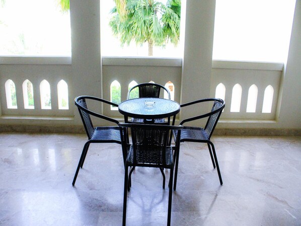 Un patio couvert avec une table ronde et quatre chaises, de grandes fenêtres à cadres blancs, et un palmier visible à l’extérieur.
