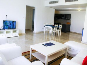 A modern living room with a white sofa, a coffee table, and a TV on a stand. There is a dining area with a table and chairs, and a kitchen in the background. - Spacious Waterfront Elegance (As Sifah)
