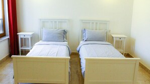Habitación doble con dos camas individuales, mesitas de noche blancas y una ventana con cortinas rojas.