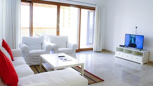 Una sala de estar con muebles blancos, televisión y puerta corrediza de vidrio al exterior.