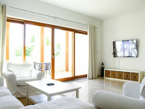 A modern living room with a white sofa, a white coffee table, a TV mounted on the wall, and a sliding glass door leading to a balcony. - Spacious 3-bedroom Luxury Living (As Sifah)