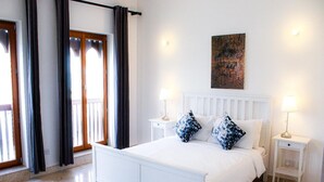 A white bed with two pillows, a nightstand with a lamp, a painting on the wall, and a balcony with curtains. - Elegant Coastal Living (As Sifah)