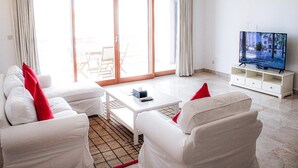 A living room with a sofa set, a coffee table, a TV on a stand, and a sliding glass door leading to an outdoor area. - Elegant Coastal Living (As Sifah)