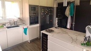 Eine moderne Küche mit weißen Schränken, einem eingebauten Backofen, einer Mikrowelle und einem Kühlschrank. Hängelampen und ein Fenster mit Vorhängen sind vorhanden.
