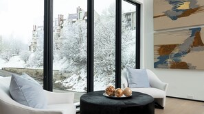 Ein modernes Zimmer mit großen Fenstern, einem runden schwarzen Ottomanen, zwei weißen Sesseln und einem Gemälde an der Wand.