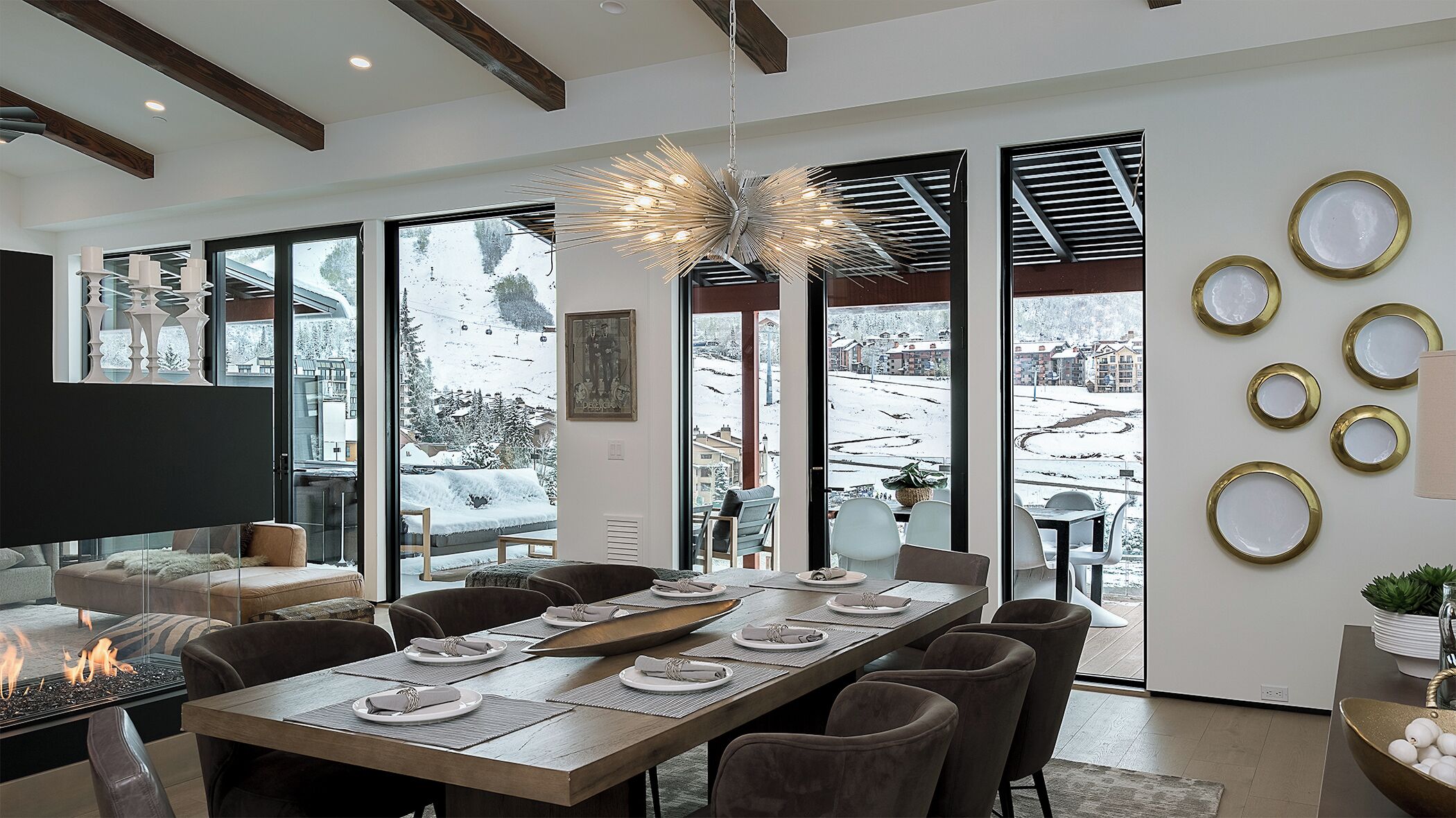 A modern dining area with a large table set for six, large windows with a snowy view, and a unique ceiling light fixture.