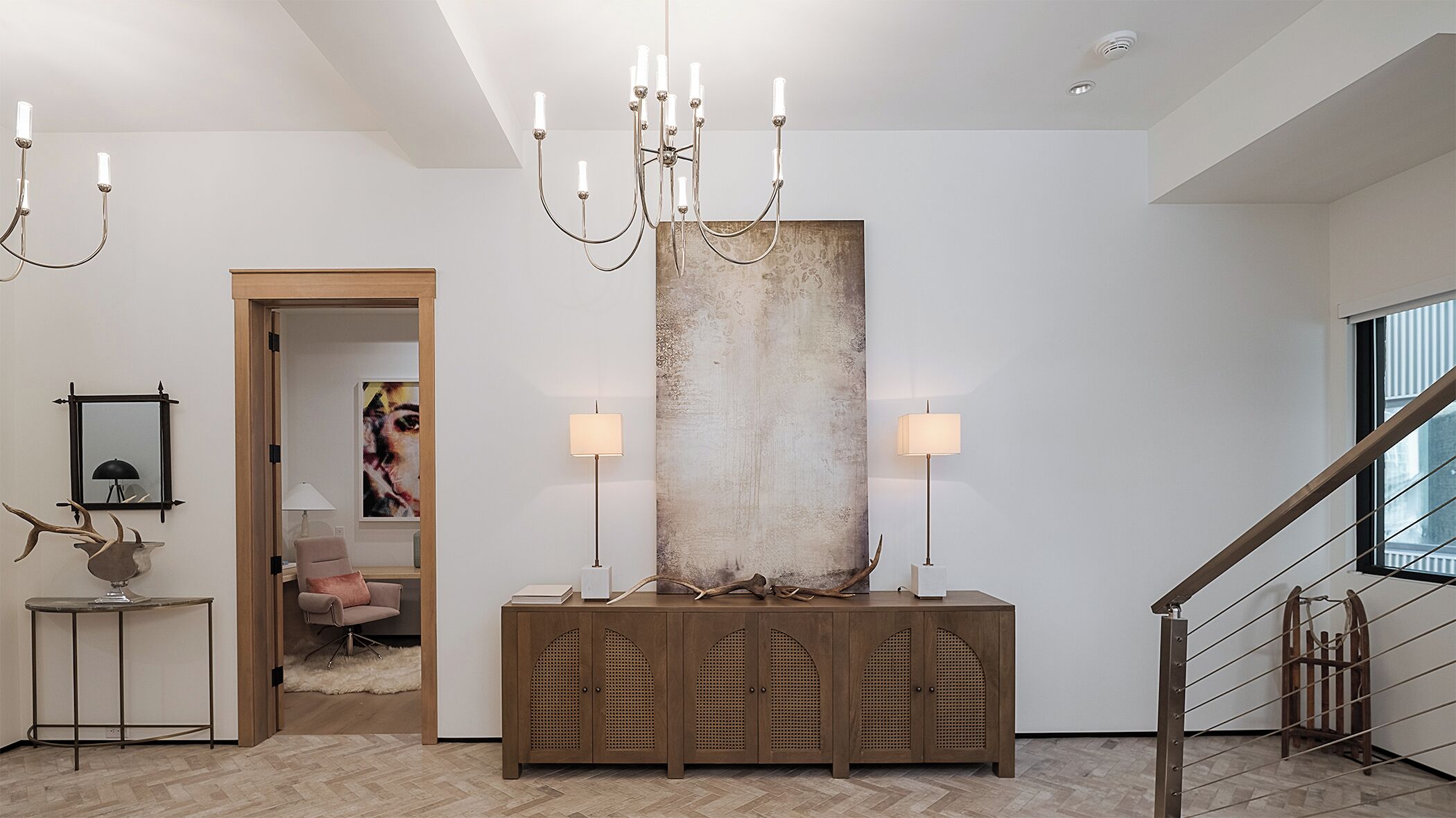 A modern interior with a wooden console, a large abstract painting, a chandelier, and a staircase.
