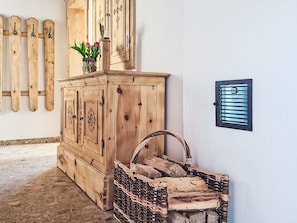 Un meuble en bois surmonté d’un vase de fleurs, un panier en osier rempli de bûches et une grille de ventilation fixée au mur.