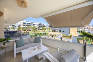 En balkon med sofa, bord og stole, med udsigt over en swimmingpool og boligbygninger.