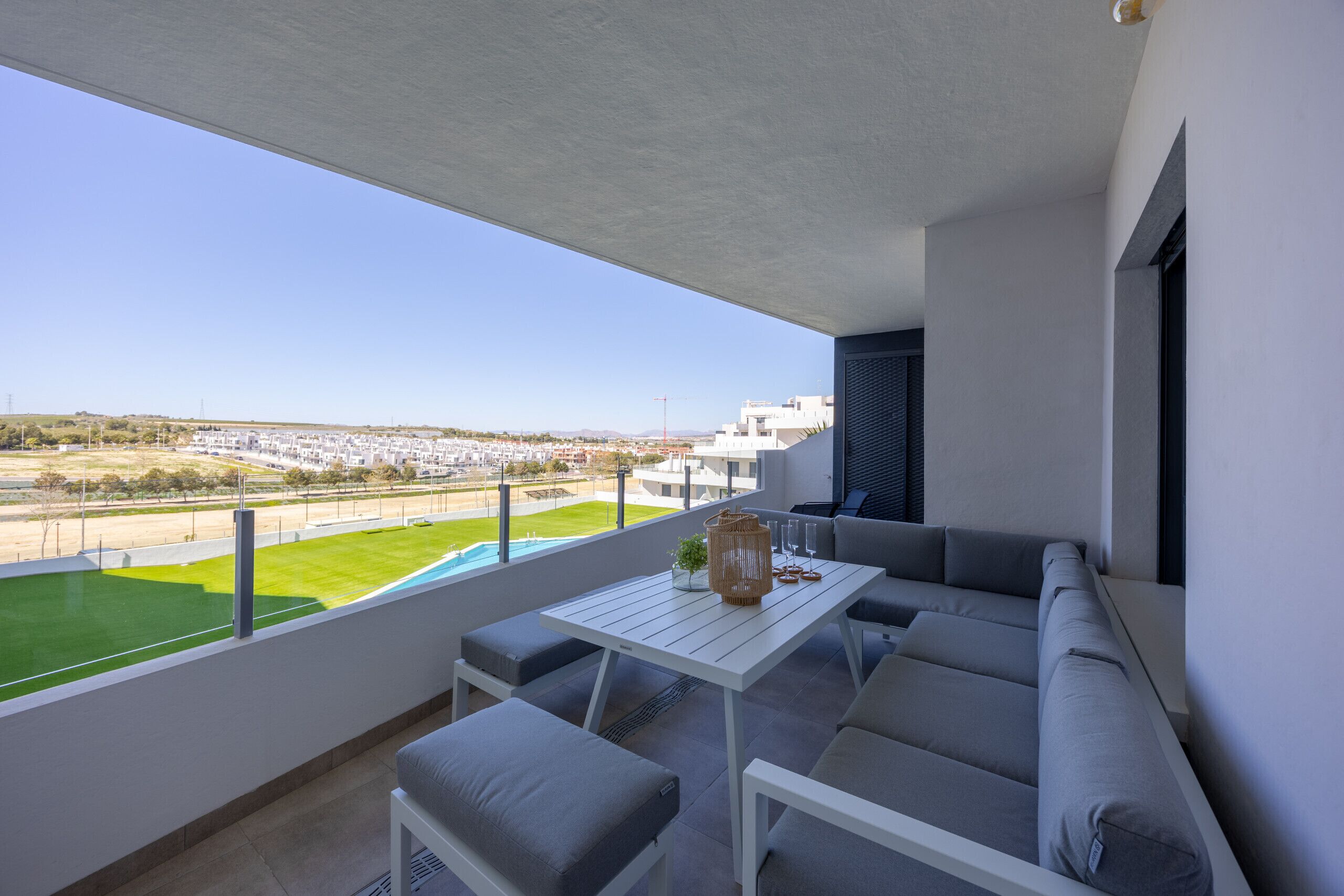 Un balcón con mesa blanca, sofá gris y vistas a un campo verde y edificios.