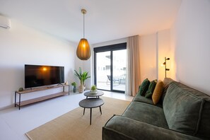 Ein modernes Wohnzimmer mit einem Flachbildfernseher, einem grünen Sofa, einem kleinen runden Tisch und einem großen Fenster.