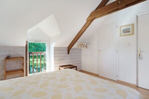 Une chambre avec un plafond en pente, un lit recouvert d’une couvre-lit à motifs de feuilles, un banc en bois, une petite table et une porte ornée d’un tableau encadré.
