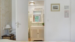 A bedroom with a bed, a bedside table with a lamp, a bathroom with a sink and mirror, and a door leading to another room. - Guest Room "La Souricière - Le Puits" with Shared Terrace, Garden & Wi-Fi (Cancale)