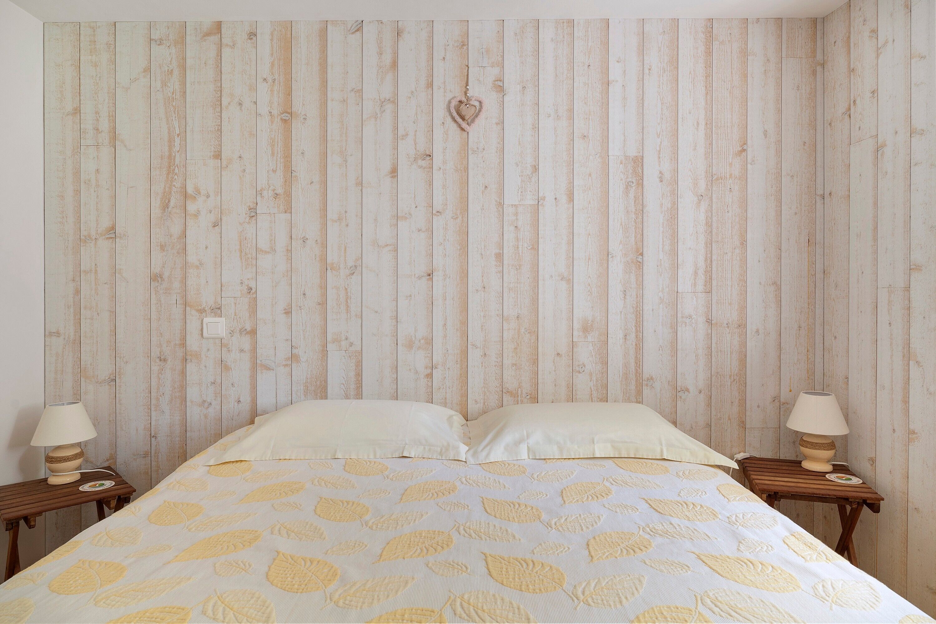 Ein Schlafzimmer mit einer Wand aus Holzleisten, einem Bett mit Bettdecke mit Blattmuster, zwei Nachttischen mit Lampen und einer herzförmigen Dekoration an der Wand.