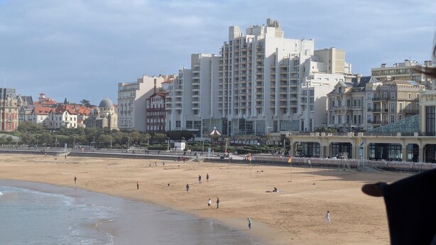 Ein Strand mit Menschen, die entlanggehen, ein sandiger Uferbereich und ein großes Gebäude im Hintergrund.