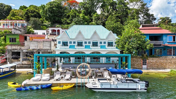 Une propriété située au bord de l’eau, comprenant une grande maison, un espace extérieur aménagé avec un coin salon couvert et plusieurs bateaux.