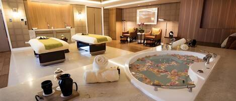 A modern hotel room with a large bathtub, two beds, a sitting area with chairs, and a small table with a vase of flowers.