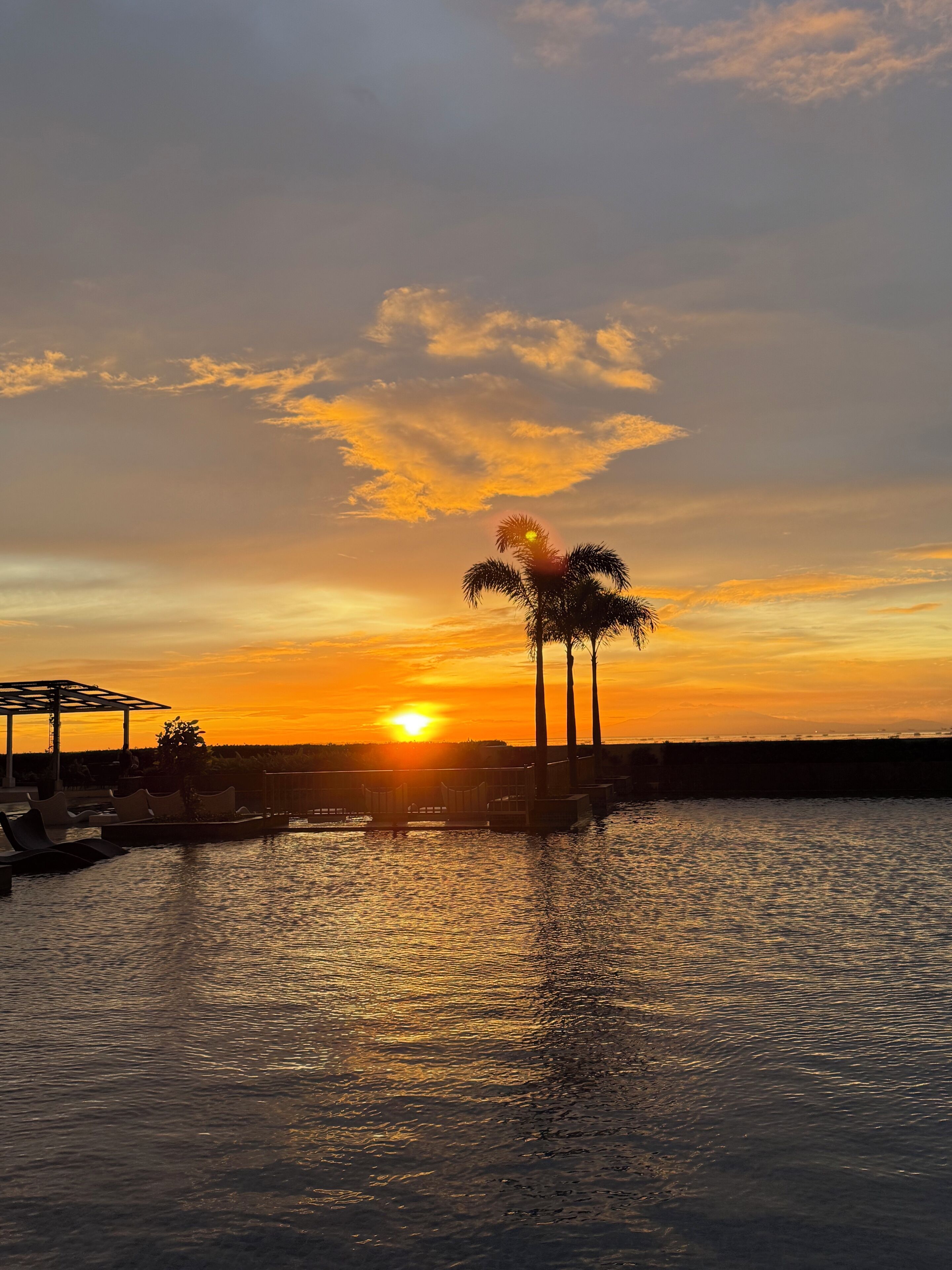 Um pôr do sol sobre a água, com palmeiras e um gazebo.