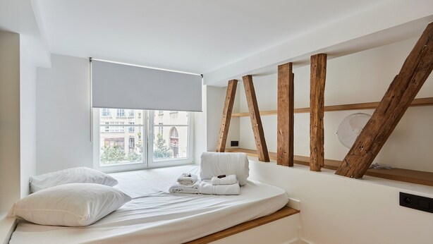 A bedroom with a bed, white bedding, a window with a blind, wooden beams, and a view of buildings outside. - Charming Duplex Loft - Paris City Center (Paris)