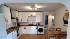 A compact kitchen with white cabinets, a built-in washing machine, and a dining area with wooden chairs and a table. - Lovely bungalow, sunny garden (Edinburgh)