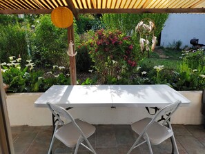A covered patio with a white table and two chairs, surrounded by lush greenery and flowering plants. - Renovated house 6 pers - Enclosed garden, terrace - 15 min from the Vendée beaches (Curzon)