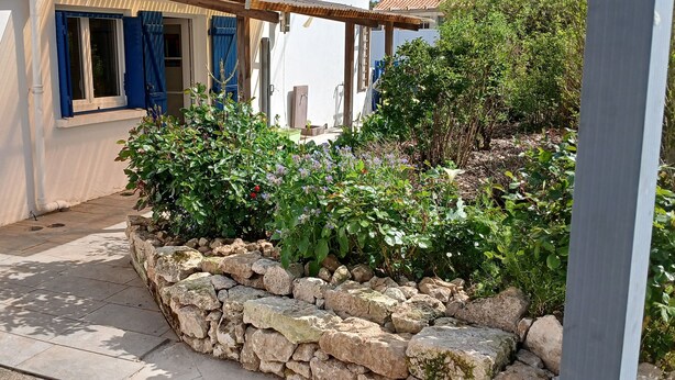 En stenplantere med grønt, en hvid bygning med blå skodder, og en pergola.